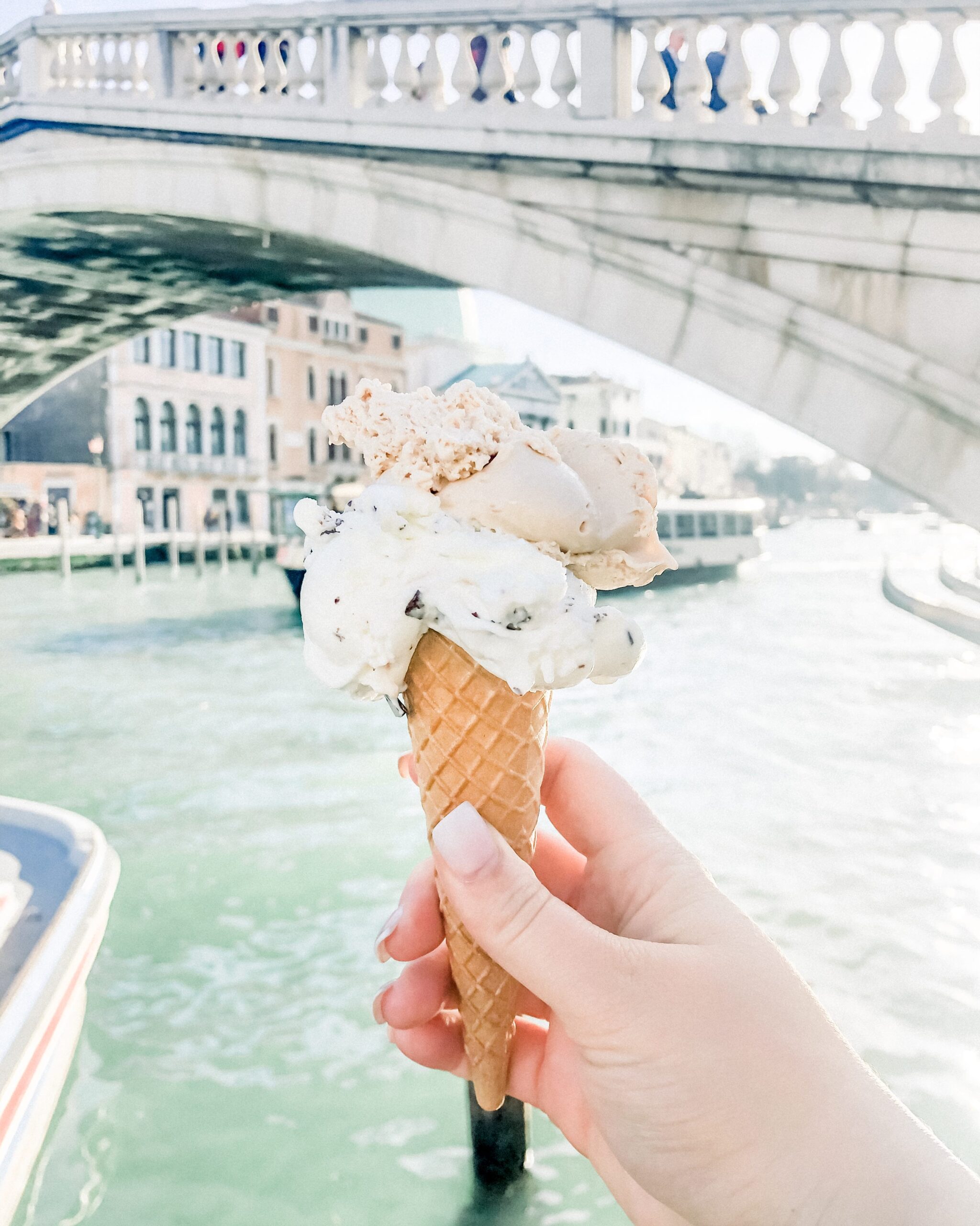 Woman's hand holding an ice cream cone with two scoops of gelato in front of the Rialto Bridge in Venice